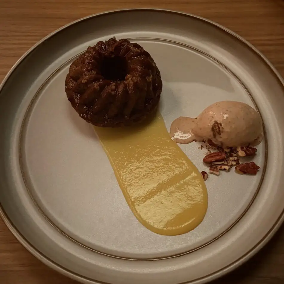 Dessert plate featuring a brown bundt cake with yellow sauce, pecan garnish, and a side scoop of ice cream