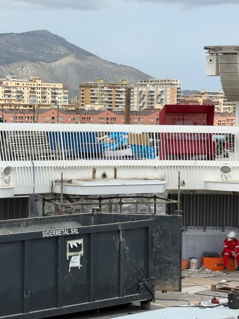 Construction site on a dock with a worker in red, a large dumpster, and city buildings in the background