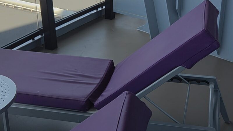 Balcony of a Virgin Voyages cruise ship cabin with purple lounge chairs and a table, overlooking a cityscape.