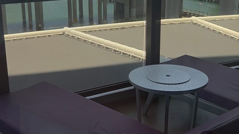 Balcony of a Virgin Voyages cruise ship cabin with two lounge chairs and a small round table, overlooking a waterway and cityscape.