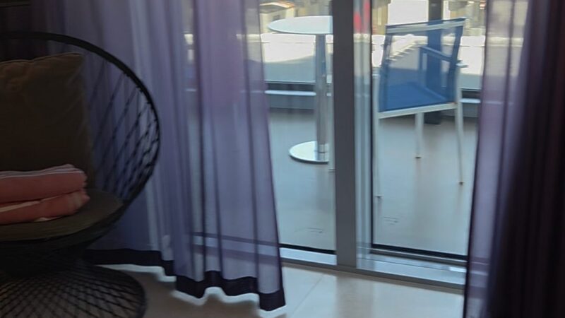 Virgin Voyages cruise ship cabin with a view of a balcony through sheer purple curtains, featuring a chair and table outside.