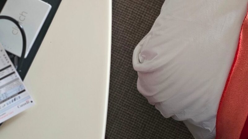 View of a Virgin Voyages cruise ship cabin showing part of a bed with white and red linens, a side table with items, and a person’s feet on a carpeted floor.