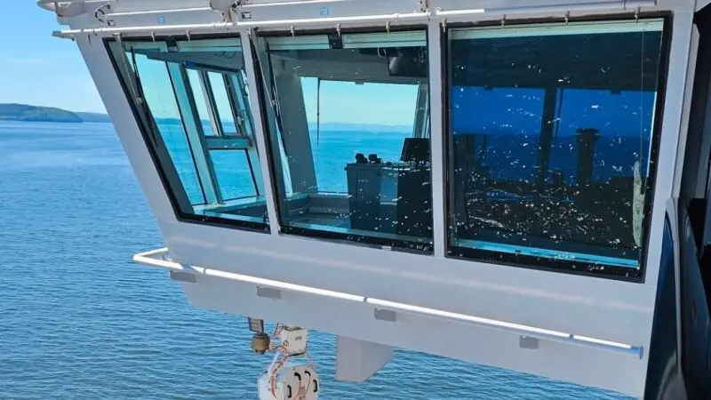 The image shows the exterior of a Virgin Voyages cruise ship with a view of the water and shoreline in the background.