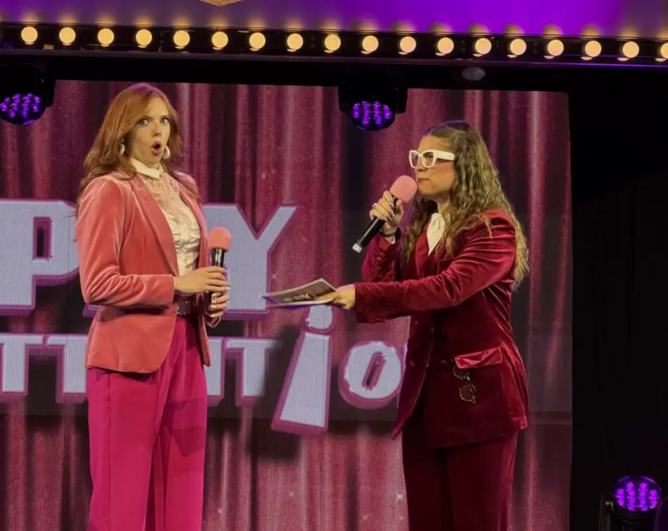 Two women perform on a stage in pink and burgundy velvet outfits holding pink microphones under purple stage lights