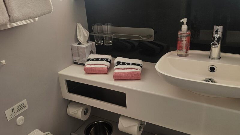 Bathroom in a Virgin Voyages cruise ship cabin, featuring a toilet, sink with a soap dispenser, towels labeled "MAKEUP," and a tissue box.