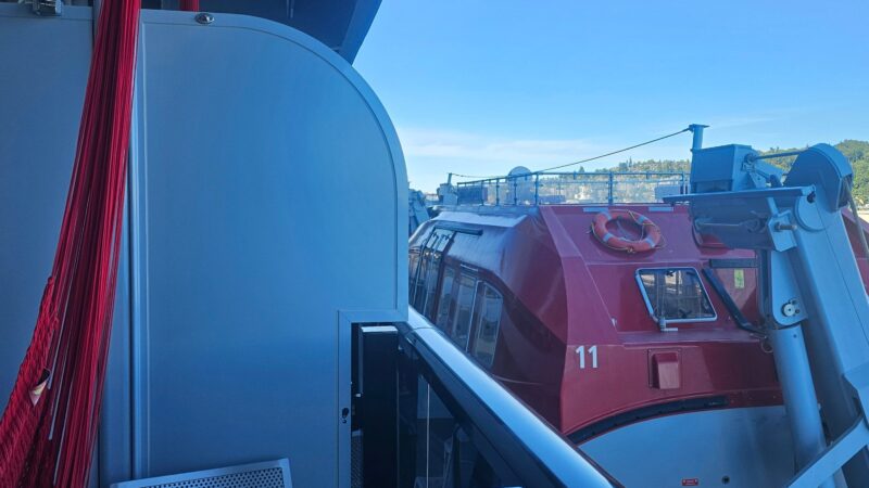 Balcony of a Virgin Voyages cruise ship cabin with a red hammock, metal chair, and lifeboat view.