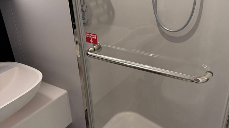 Shower area and sink in a Virgin Voyages cruise ship cabin, featuring a glass door, handrail, and toiletries on a shelf.