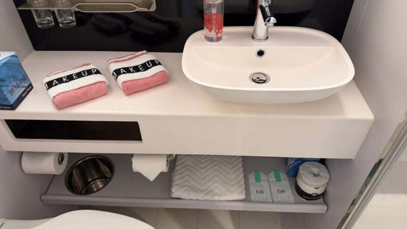 Cruise ship bathroom with a sink, faucet, soap dispenser, and towels labeled “Makeup.” Shelves hold a tissue box, extra toilet paper, and toiletries.