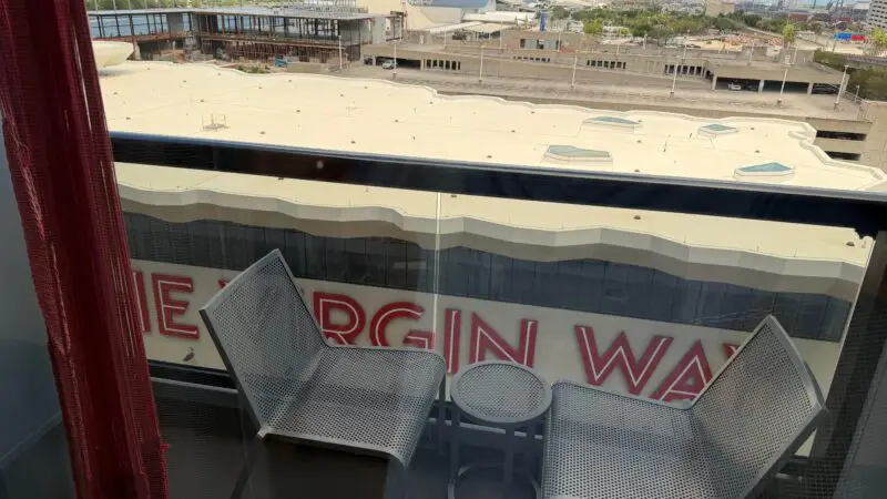 View from a Virgin Voyages cruise ship cabin showing a balcony with two chairs and a small table, overlooking the port and other cruise ships.