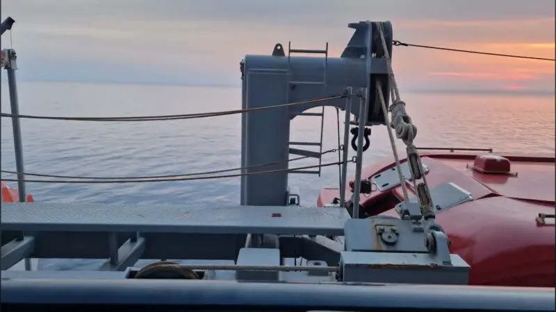 View from a Virgin Voyages cruise ship balcony, showing a lifeboat and ocean at sunset.
