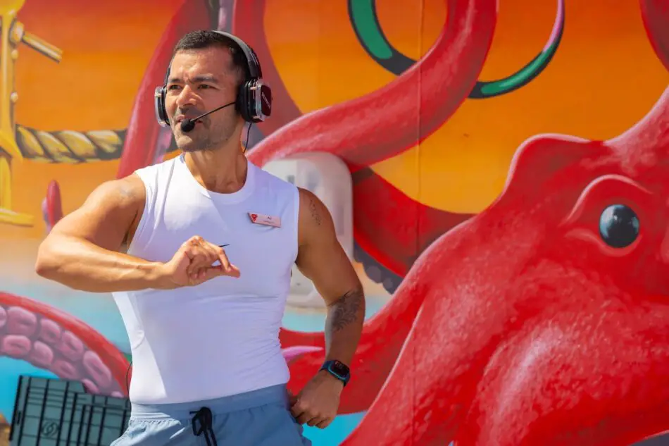 Male fitness trainer wearing headset and white sleeveless shirt, instructing a workout in front of a bright mural