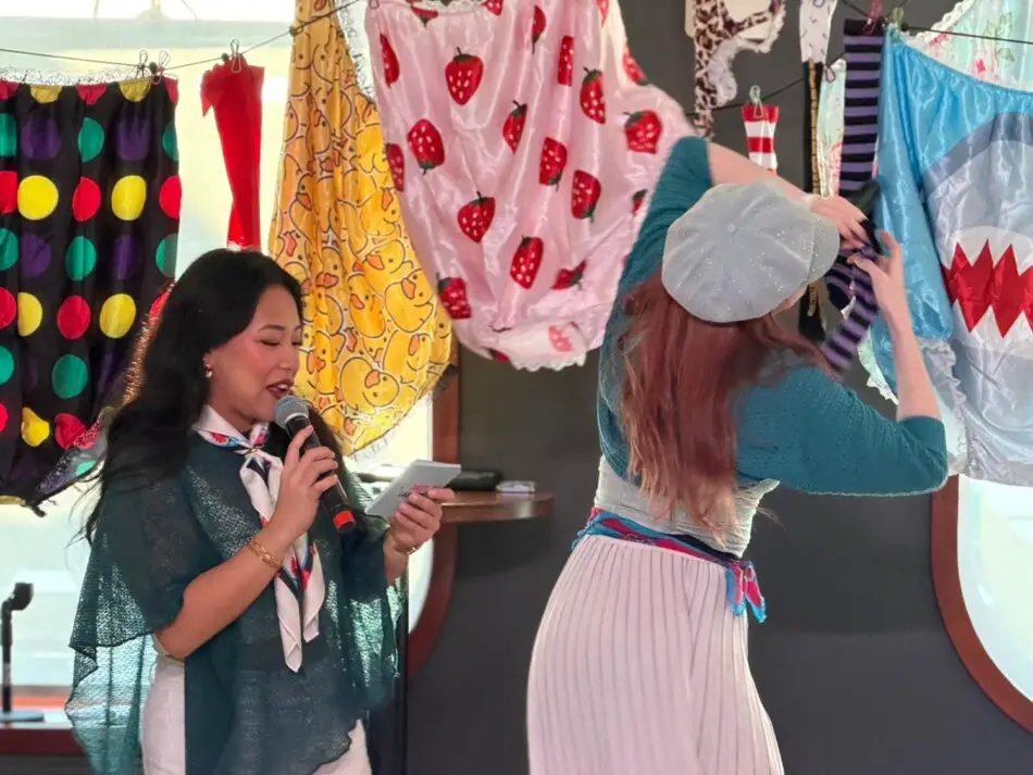 Woman speaks into microphone on stage while another adjusts colorful fabrics