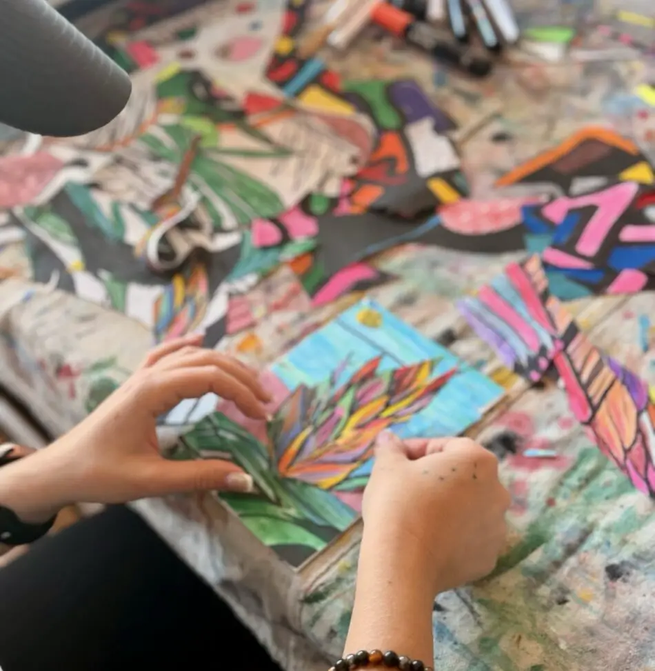 Two hands assemble a colorful paper collage on a paint-splattered desk