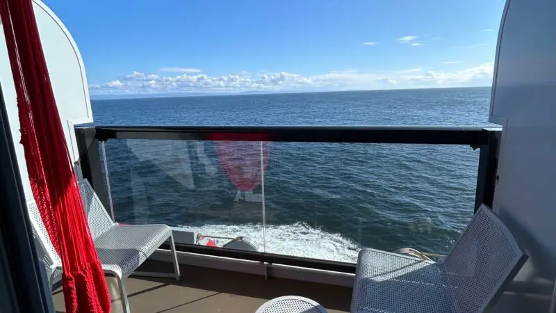 Balcony of a Virgin Voyages cruise ship cabin with two chairs, a small table, and an ocean view under a clear blue sky.