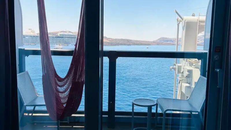 Cruise ship cabin balcony with two white chairs, a small table, and a red hammock facing the ocean.