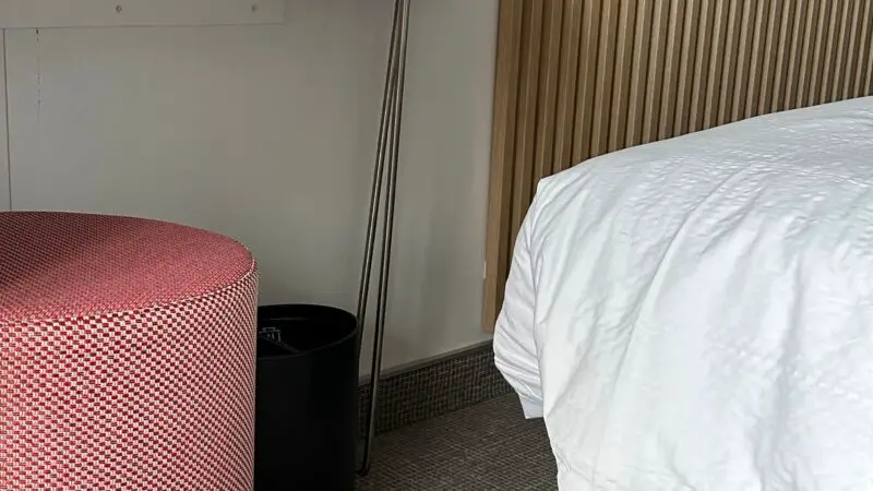 Interior of a Virgin Voyages cruise ship cabin featuring a white bed, a round pink stool, and a wooden slat wall.
