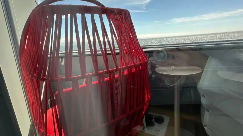 Balcony view of a Virgin Voyages cruise ship cabin featuring a red hanging chair and a table, overlooking the ocean with a clear blue sky.