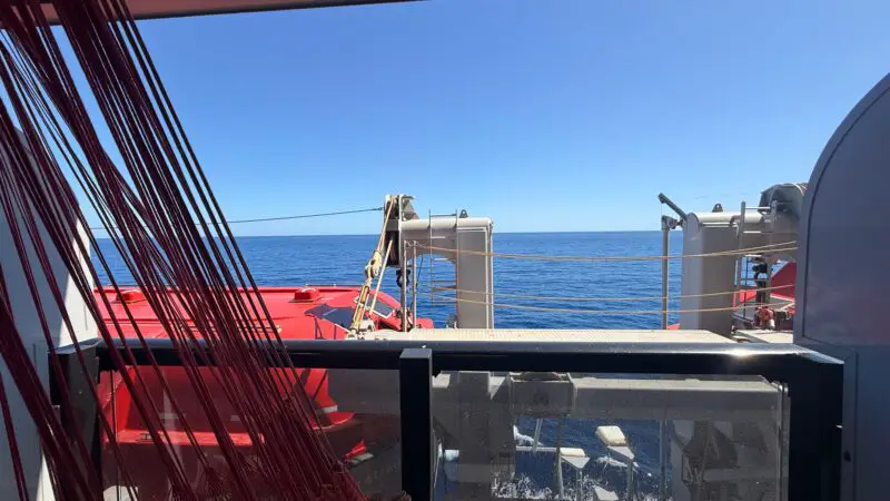 View from a Virgin Voyages cruise ship cabin balcony with red hammock, looking out at the calm sea and blue sky.