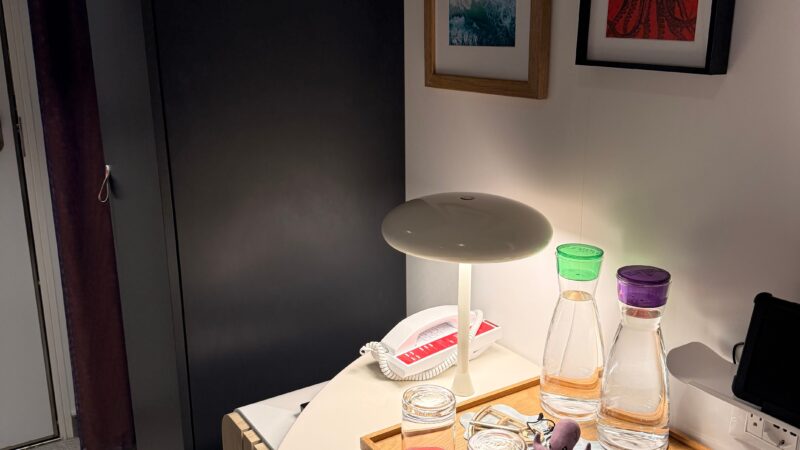 Virgin Voyages cruise ship cabin with a table featuring water carafes, glasses, and a phone. Wall art includes an ocean scene and an octopus.