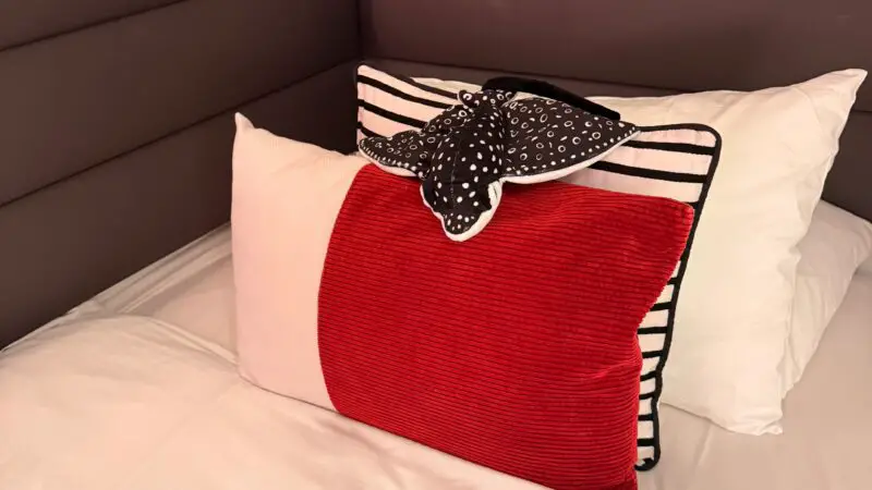 Virgin Voyages cruise ship cabin bed with white linens, a red pillow, and a decorative towel folded into a shape.