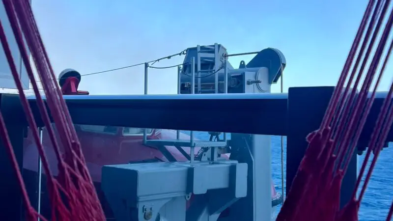 View from a Virgin Voyages cruise ship cabin balcony, showing feet resting in a red hammock with the ocean and ship railings in the background.