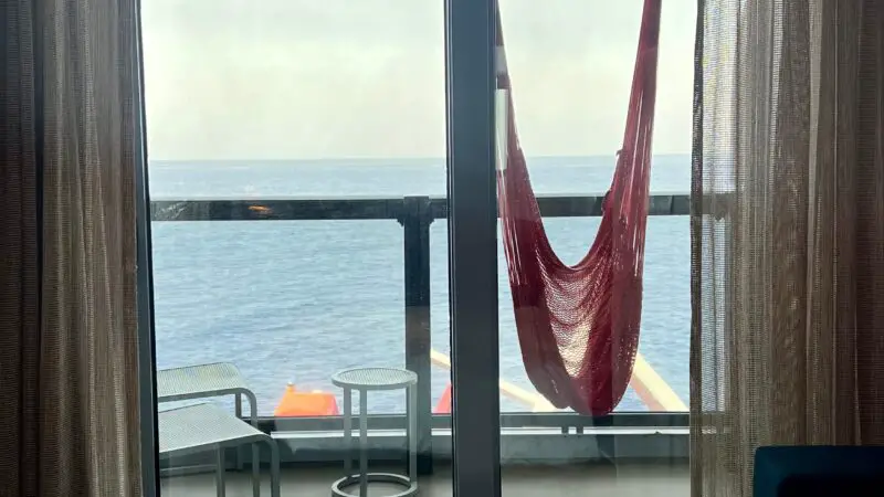 Virgin Voyages cruise ship cabin with a bed facing a balcony. A red hammock and seating are visible outside with an ocean view.