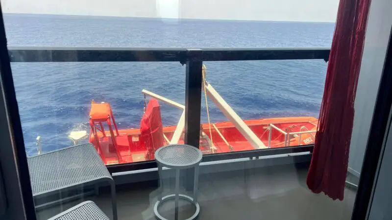 View from a Virgin Voyages cruise ship balcony cabin, showing a small table, chairs, and ocean scenery beyond a glass railing.