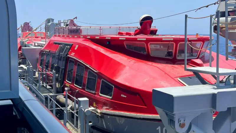 The image shows a bright red lifeboat positioned on the deck of a Virgin Voyages cruise ship under a clear blue sky.