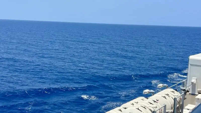 View from a Virgin Voyages cruise ship balcony, showing lifeboats and a red safety boat with the ocean and sky in the background.