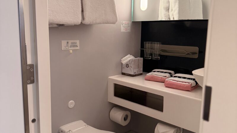 Interior of a Virgin Voyages cruise ship cabin bathroom featuring a modern toilet, white towels, a sink with toiletries, and a mirrored vanity.
