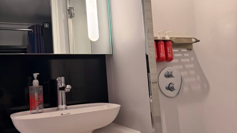 Bathroom area of a Virgin Voyages cruise ship cabin, showing a modern sink with a mirror, a shelf with red toiletry bottles, and a white wall with shower controls.