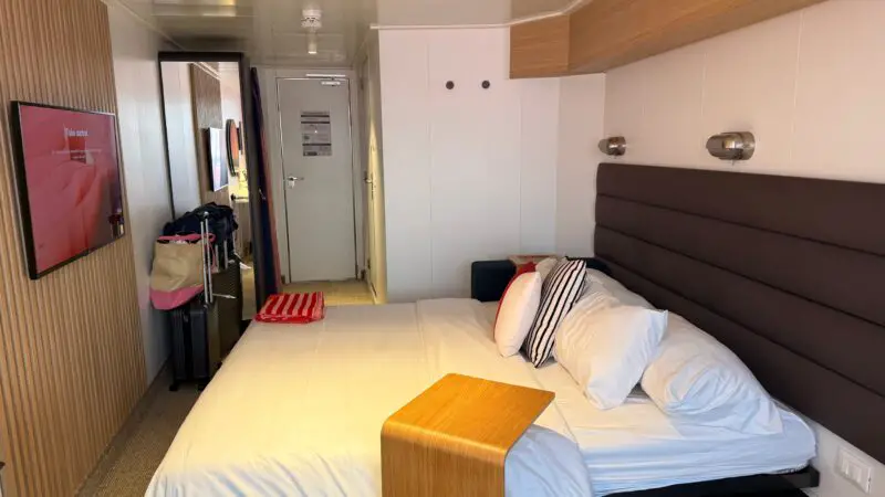 Virgin Voyages cruise ship cabin with a bed, striped pillows, a small table, wall-mounted lights, a TV, and a doorway with luggage and bags nearby.