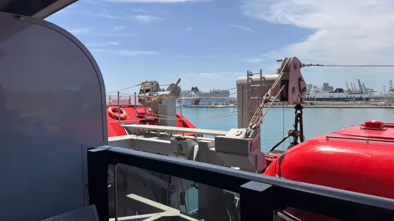 View from a Virgin Voyages cruise ship balcony, showing lifeboats and ship equipment against a backdrop of the sea and distant port.