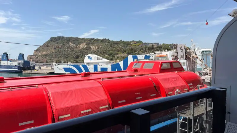 View from a Virgin Voyages cruise ship balcony overlooking a harbor with other ships, a red lifeboat in the foreground, and a hillside in the background under a blue sky.