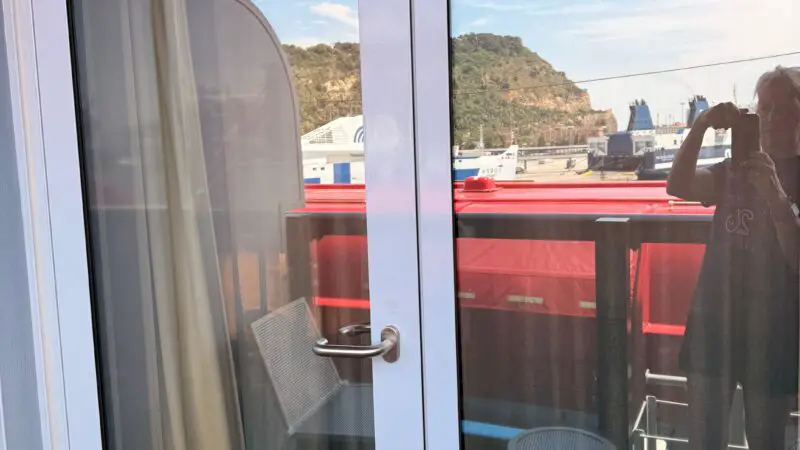 Glass door of a Virgin Voyages cruise ship cabin leading to a balcony, with a partial reflection of the surroundings visible.