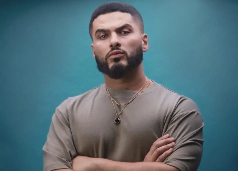 Man with a beard and necklace, arms crossed against teal background