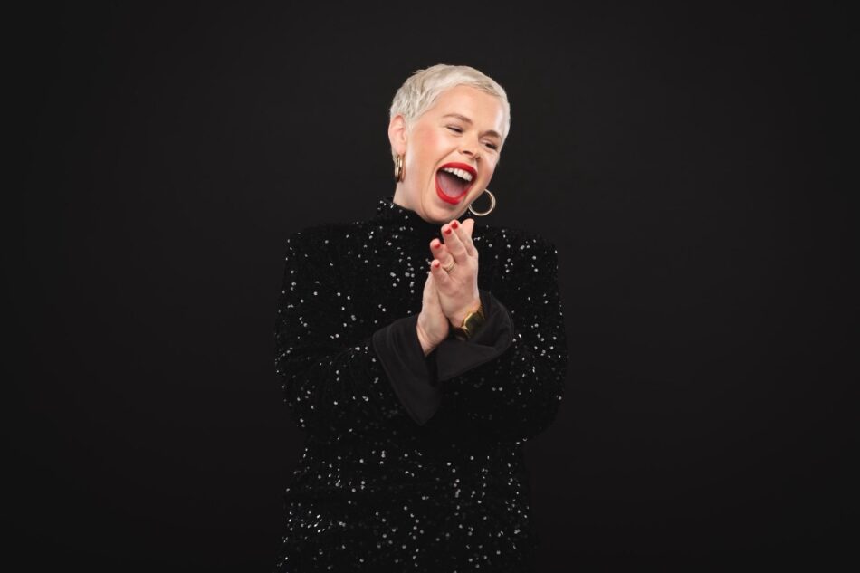 Woman with short silver hair wearing a black sequin top, smiling and clapping on a dark background
