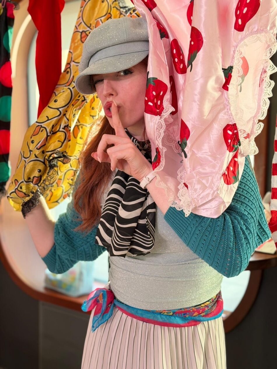Woman wearing a gray cap and teal sweater among colorful dresses in a boutique