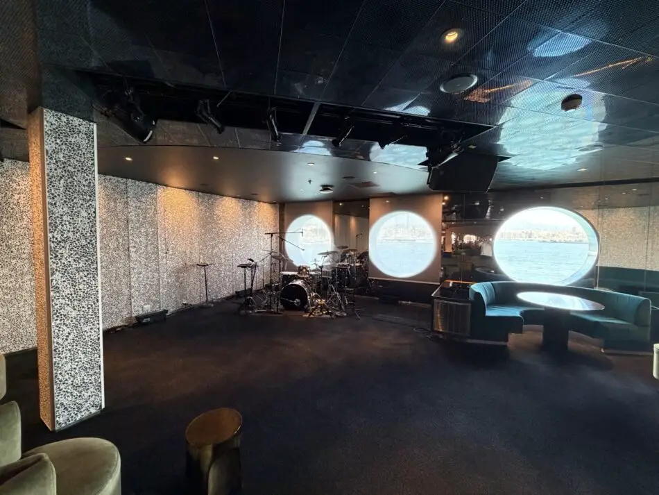 Stage with drum kit and seating area on a Virgin Voyages cruise ship, circular windows overlook the sea