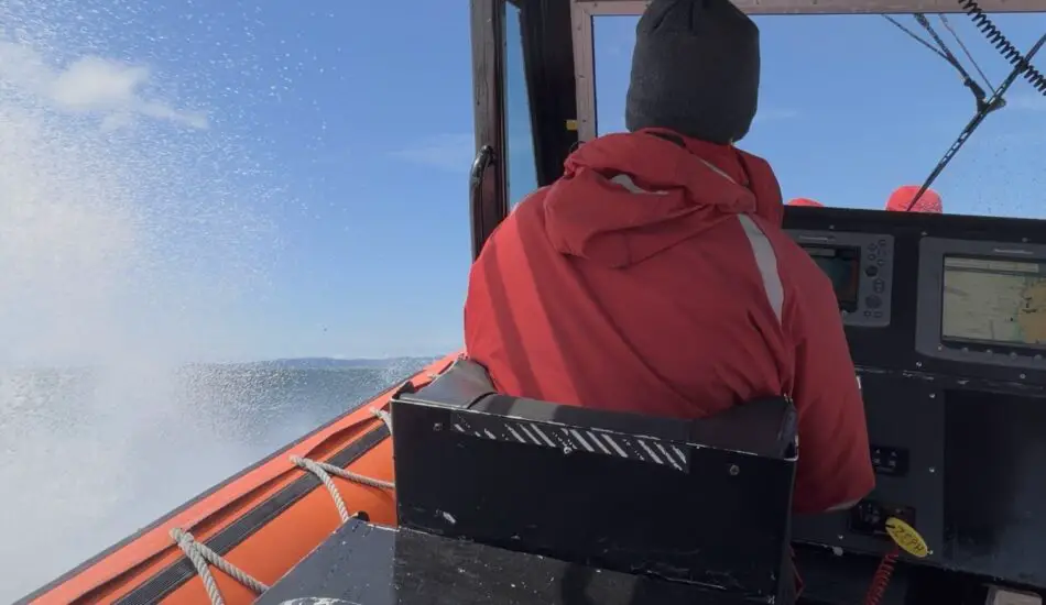 Person in red jacket and gray beanie in a boat cockpit with navigation screens and ocean spray