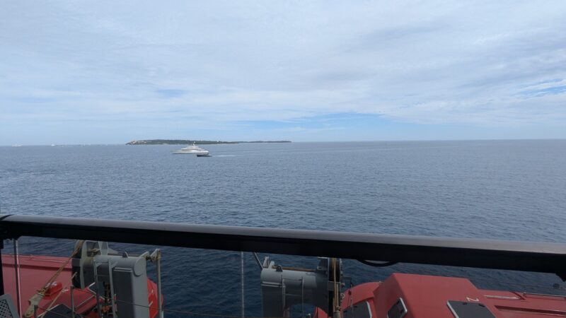 View from a Virgin Voyages cruise ship balcony overlooking the ocean, with a yacht in the distance and part of the ship's life-saving equipment visible below.