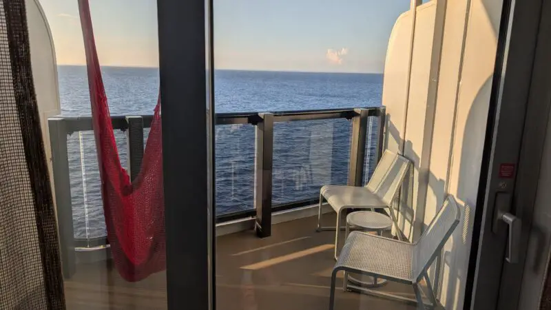 View from a Virgin Voyages cruise ship cabin balcony, featuring two chairs, a small table, and a red hammock with the ocean visible beyond the railing.