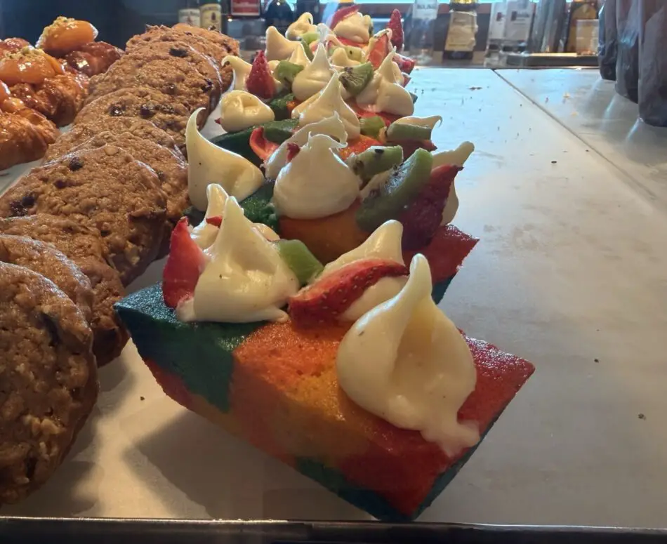 Rainbow cake slice topped with whipped cream and fruit beside cookies in a bakery display
