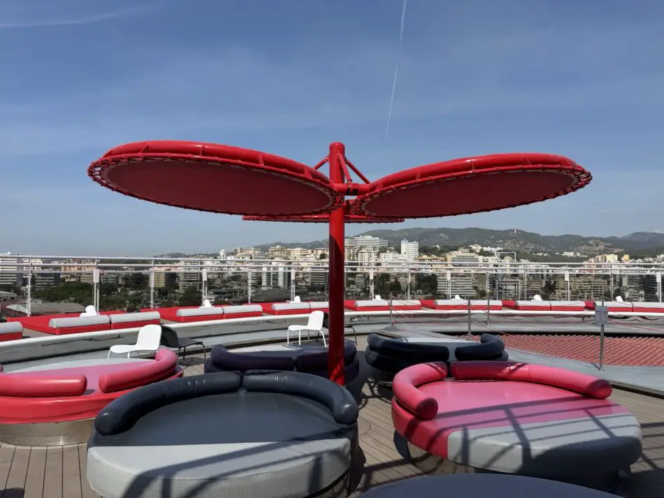 Red circular lounge pods on a Virgin Voyages cruise ship rooftop deck with city skyline