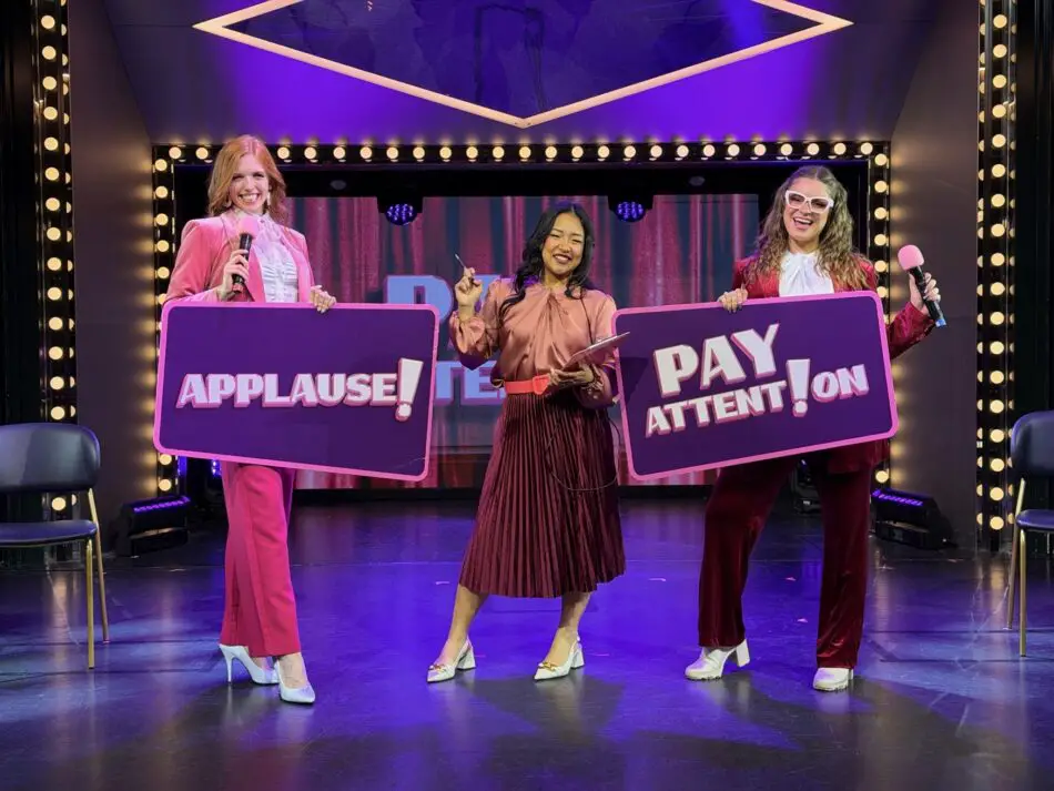 Three women on a stage holding large purple signs reading APPLAUSE! and PAY ATTENTION!