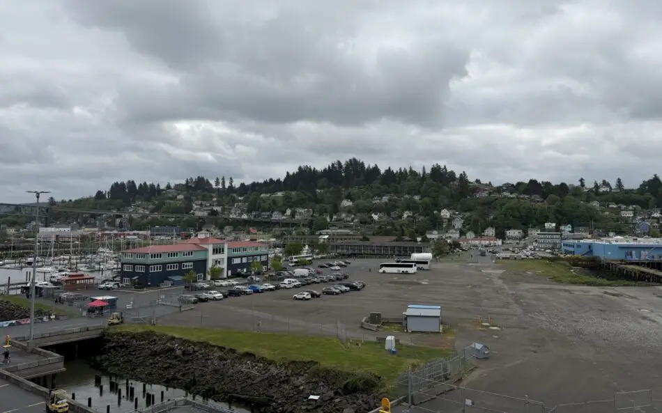Coastal harbor with boats, a large parking lot, and hillside houses under a cloudy sky
