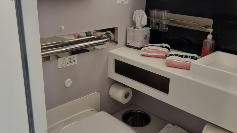 Cruise ship cabin bathroom with a wall-mounted toilet, towels, tissue box, glasses, soap dispenser, and a mirror above a counter.