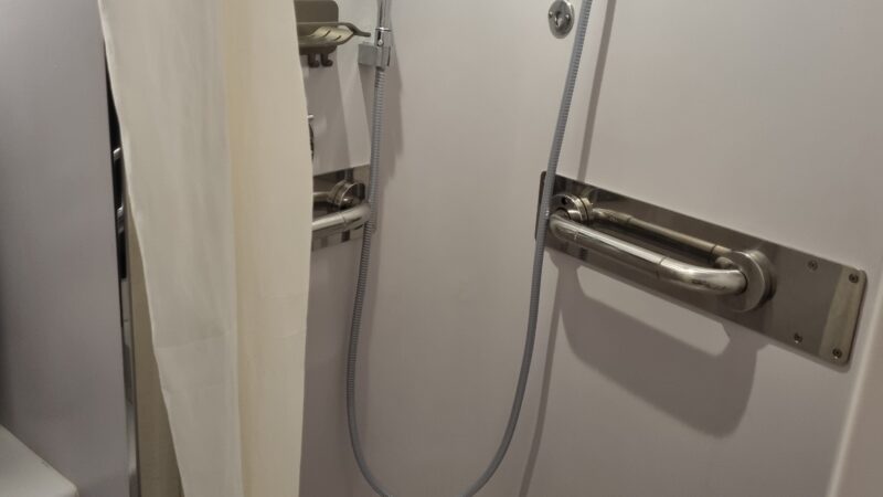 Cruise ship cabin shower with handheld showerhead, grab bar, and white curtain.