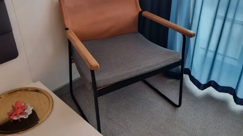 A Virgin Voyages cruise ship cabin featuring a leather and fabric chair next to a curtain-covered window, with a small side table holding a decorative tray and a folded towel.