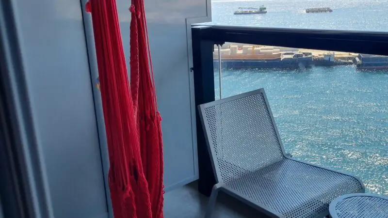 Balcony of a Virgin Voyages cruise ship cabin featuring a red hammock, metal chair, and small table, with a view of the sea and distant mountains.